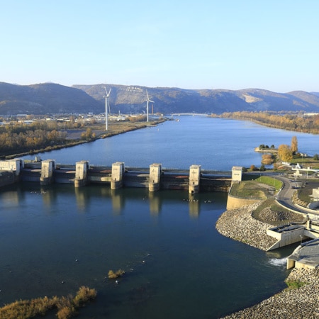 Photographie aérienne montrant un barrage, le canal de navigation et les infrastructures associées. Le Rhône traverse un paysage composé de zones naturelles et d’ouvrages hydrauliques.