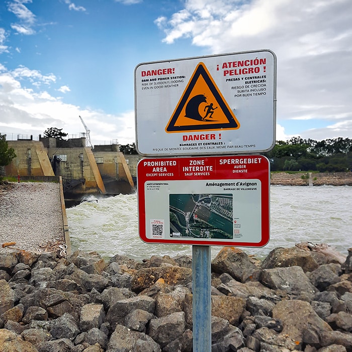 Un panneau d’avertissement est implanté sur une berge rocheuse devant un barrage. Il indique les risques liés aux variations du débit et aux montées soudaines des eaux dans cette zone réglementée.