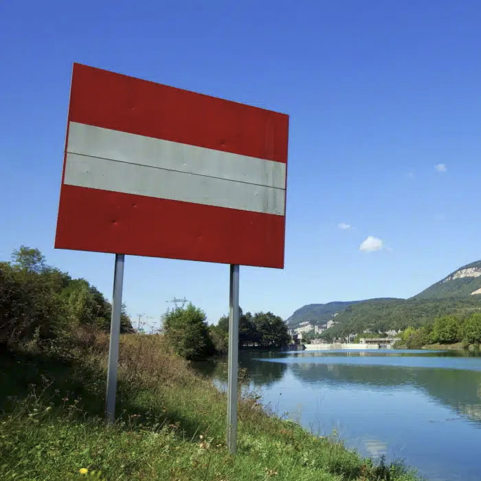 Panneau d’interdiction rouge et blanc installé au bord d’un cours d’eau, symbolisant la sécurité, le contrôle d’accès et la gestion des risques en milieu industriel, avec des collines et un ciel dégagé en arrière-plan.